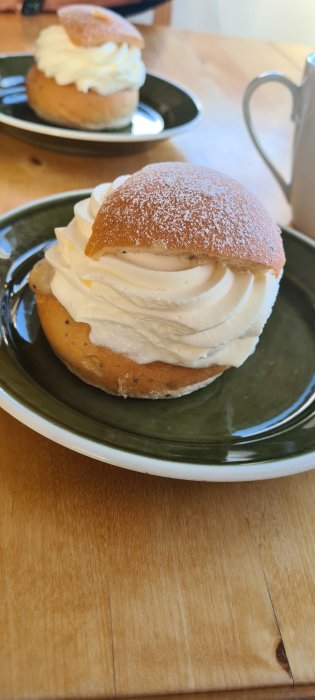 Svensk semla med vispad grädde på ett grönt fat, ett kaffekoppshandtag syns i bakgrunden.