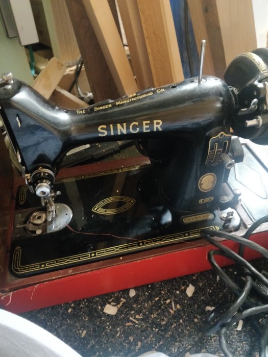 Gammal Singer symaskin i svart med detaljer av guld, står på en röd kant. Saknar täcklock över undertråden.