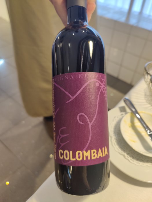 En hand håller en flaska vitt vin med etiketten "Colombaia Vigna Nuova" vid ett bord med en tom desserttallrik.