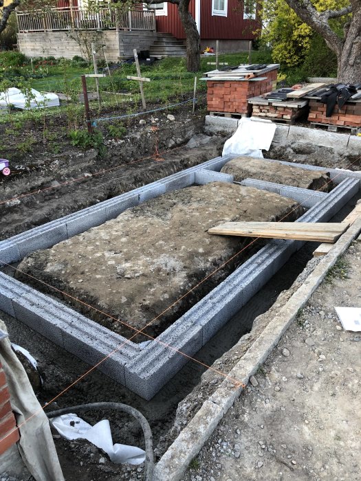 Grundläggning för växthus med betongblock och armering på en grävd tomt med odlingsland i bakgrunden.