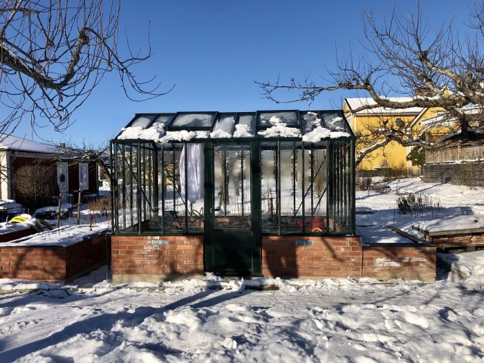 Växthus med bas av murade tegelstenar i snöig trädgård, klar för tegelgolvinstallation.