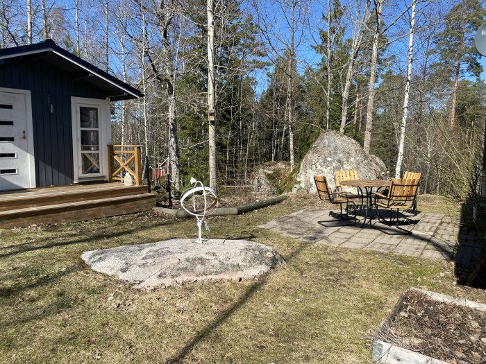 Trädgård med befintligt trädeck, stuga, och utemöbler på stenplattor omgivna av skog och sten.