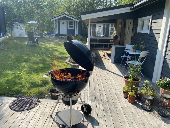 Grill på ett trädäck vid en stuga med utemöbler och planteringar, grön gräsmatta och förråd i bakgrunden.