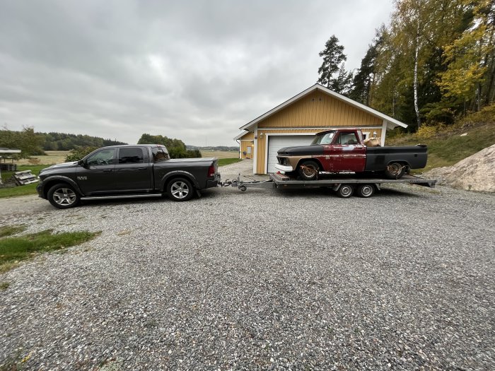 En modern pickup drar ett släp med en klassisk bil, förmodligen en Chevrolet C10, framför ett garage.