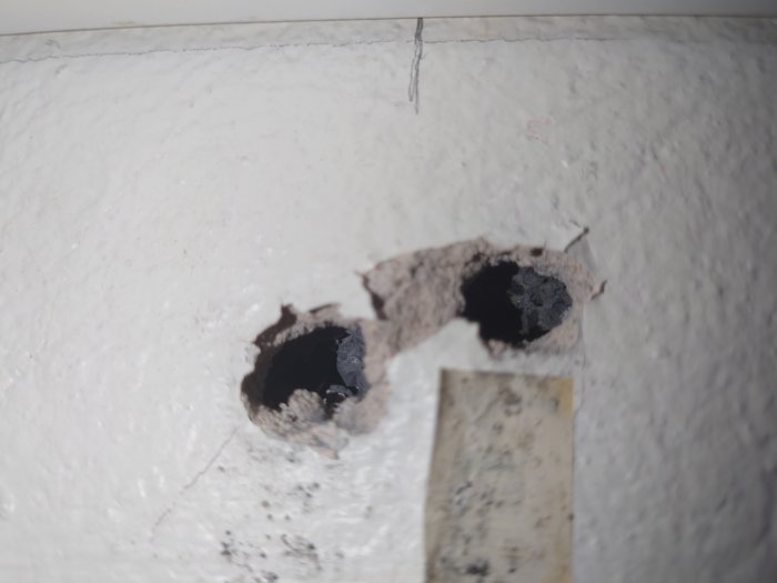 Två urgröpta hål i en vit vägg med spår av tidigare borrning och fragmenterad kant.