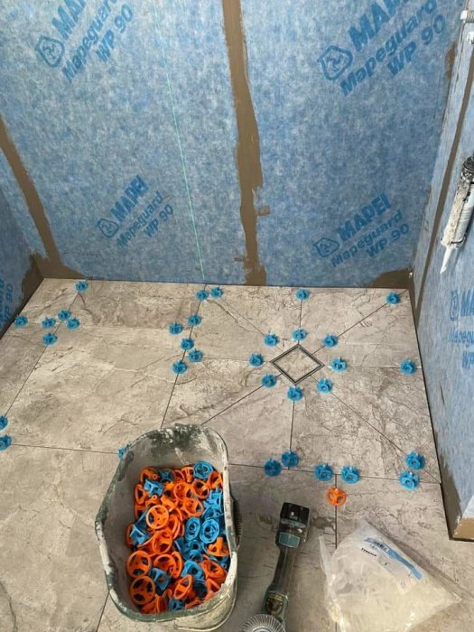 Badrum under konstruktion med stora kakelplattor, blå och orange nivelleringssystem och vattentätning på väggarna.
