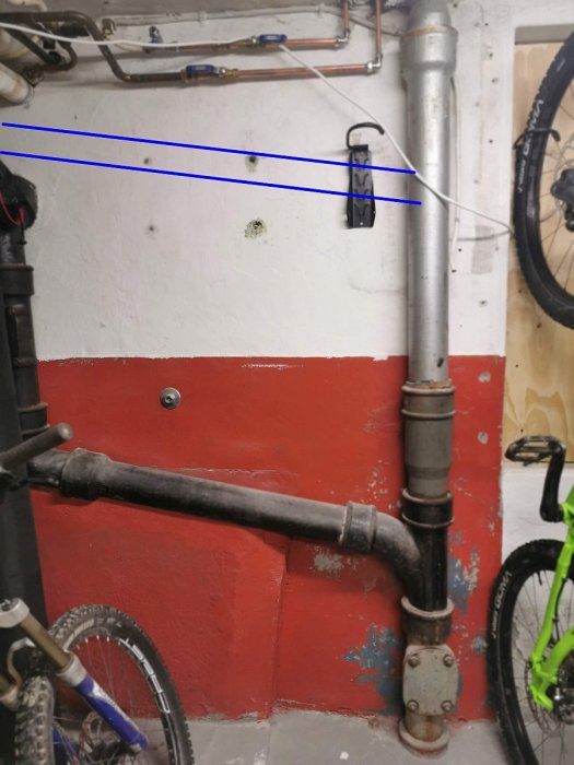 Gjutjärnsrör och plastavlopp kopplade med blå markerad skarv, nära en cykel i ett rödmålat källarutrymme.