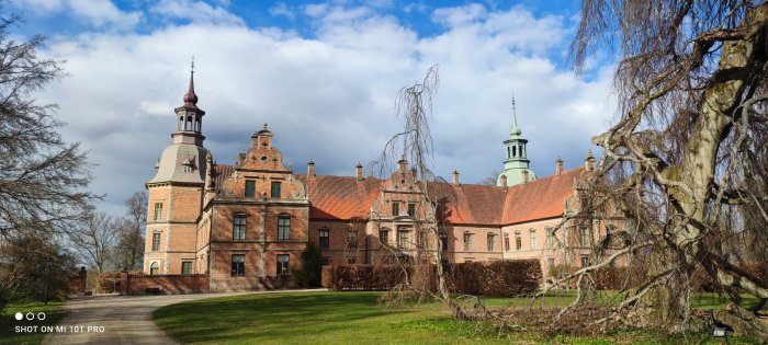 Historiskt tegelstensslott med torn och spiror omgivet av grönska under en klar blå himmel.