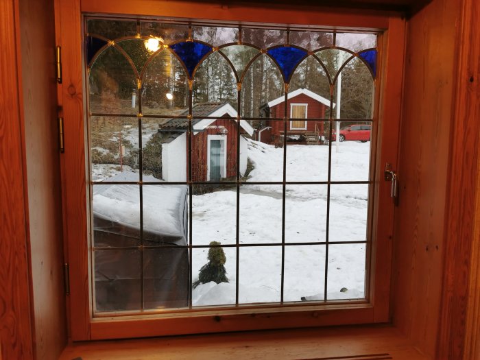 Träfönster med överbågar i färgat glas som öppnas inåt, med utsikt över snötäckt trädgård och rött hus.