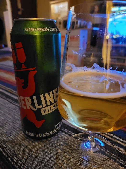 En burk Berliner Pilsner bredvid ett halvfullt glas öl på ett rutmönstrat underlag.