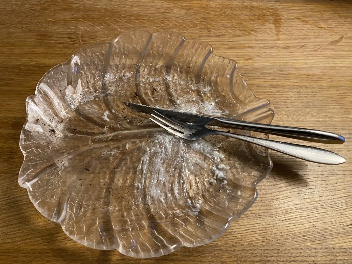 En tom, genomskinlig tallrik i form av ett blad med rester av en måltid och bestick på ett träbord.