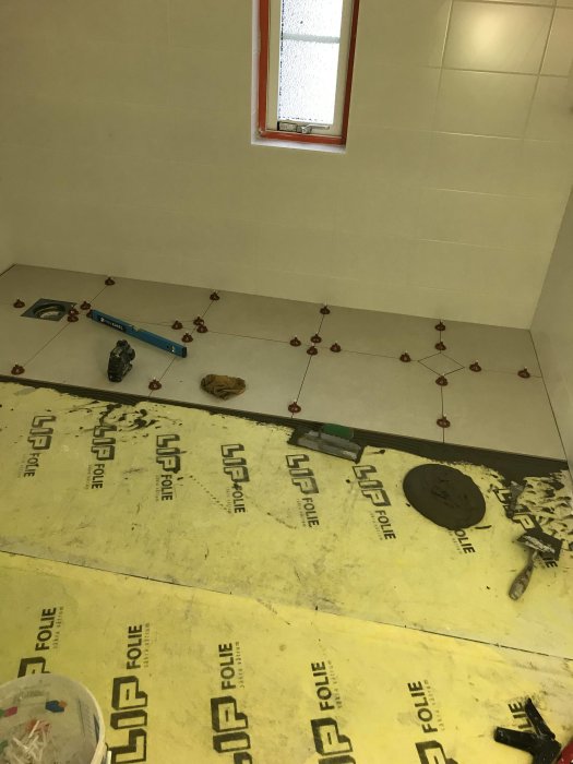 Pågående kakelsättning i ett rum med nylagt golvkakel och delvis kaklade väggar, verktyg syns på golvet.