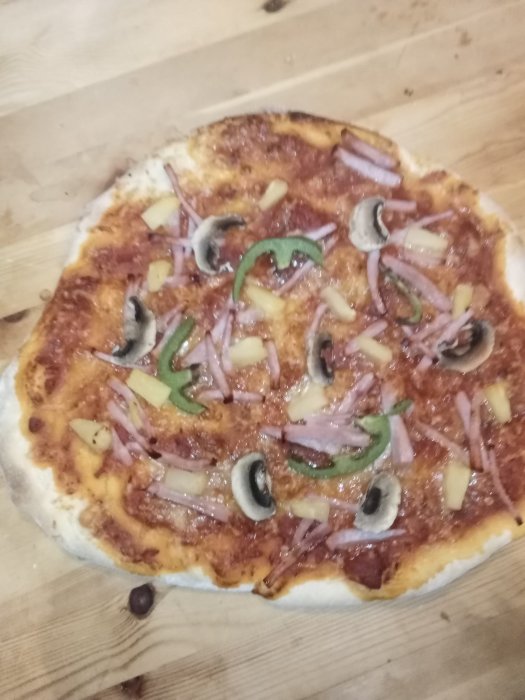 Hemlagad pizza med skinka, ananas, lök, paprika och svarta oliver på träbänk.