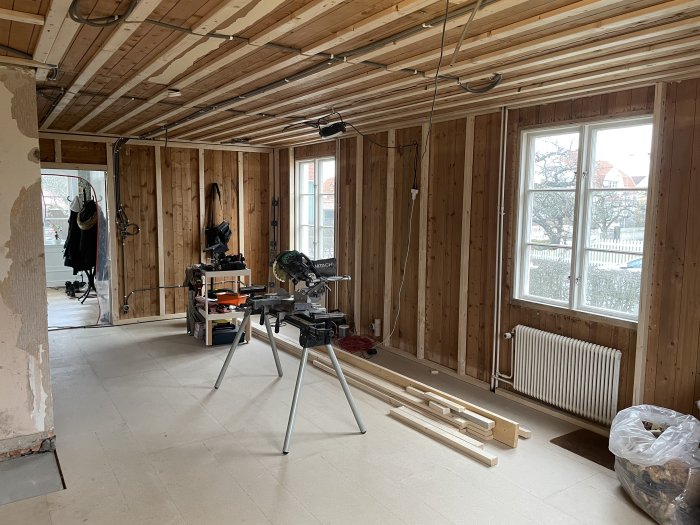 Vardagsrum under renovering med reglade väggar och tak, synliga elinstallationer och verktyg på golvet.