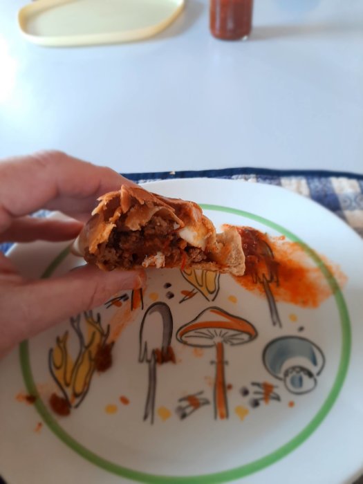 Halvöppnad chilensk empanada med fyllning i handen ovanför en tallrik med Aji-såsfläckar.