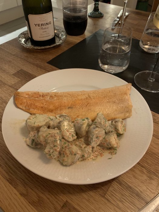 Rödingfilé med mandelpotatis i sås på tallrik, vinflaska och glas i bakgrunden.