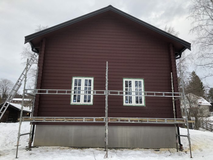 Nyinstallerade fönster på röd garagevägg med ställning, stege, och snö i förgrunden.