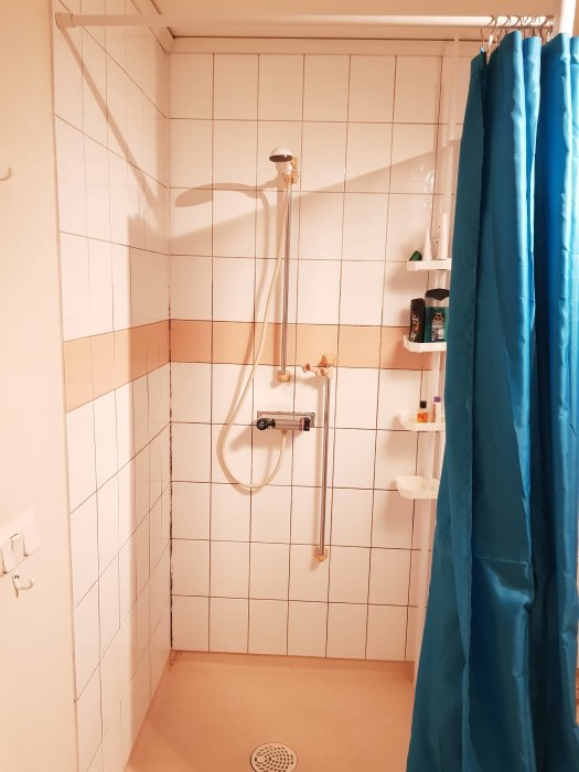 Hörnet av ett badrum med vita kakelväggar, nyinstallerad takdusch från Grohe och en blå duschdraperi.