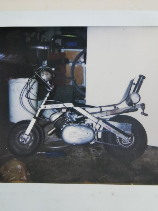 En miniatyrmotorcykel står i ett rörigt garage, bilden är en polaroid.