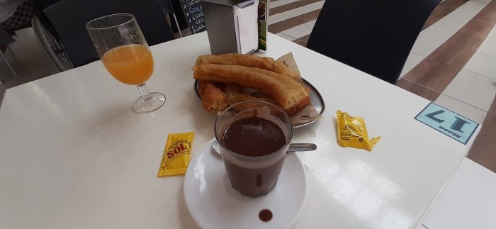 Nyfriterade churros med strösocker vid sidan av varm choklad och ett glas apelsinjuice på ett cafébord.