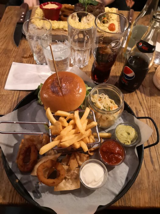 Middag på krog med hamburgare, pommes frites, lökringar och diverse såser, samt drycker på bordet.