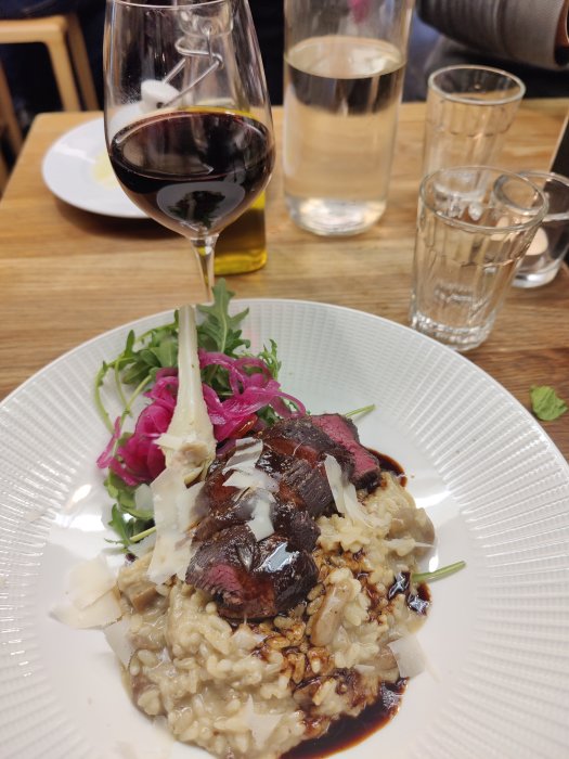 Tallrik med risotto toppad med skivor av stekt kött, rucola och hyvlad ost, vin och vattenglas i bakgrunden.