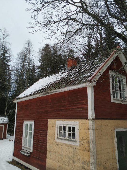 Gammal gäststuga med buktande röd yttervägg, snö på tak med ojämna tegelpannor och synlig fuktskada på nedre brädan.