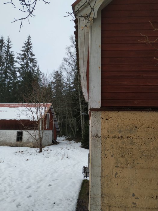Buktande yttervägg på gäststuga med skadad bräda nedanför takkanten, omgiven av snö.