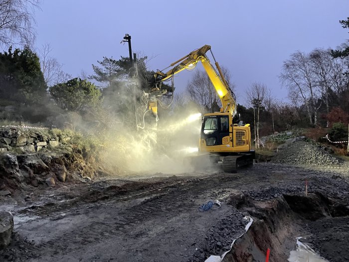 Gul grävmaskin utför markarbete och bergsschaktning med en hydraulisk spräckare i en trädgård.