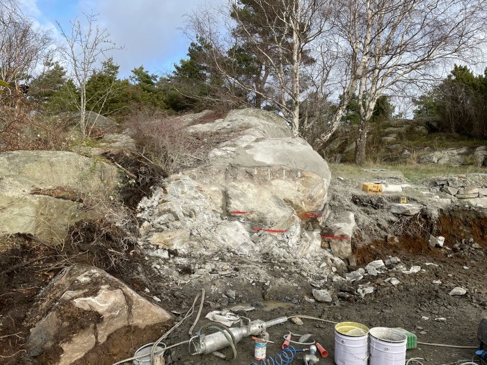 Bergskrapning på byggarbetsplats med synlig bergspräckare, hinkar, borrmaskin och skog i bakgrunden.