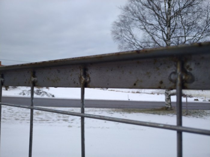 Närbild på en metalgrind med tecken på rost och slitage, med snöigt landskap i bakgrunden.