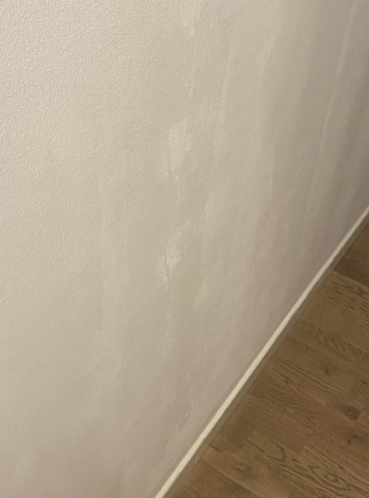 Vägg med ojämn målning och fläckar i släpande ljus, ovan trägolv.