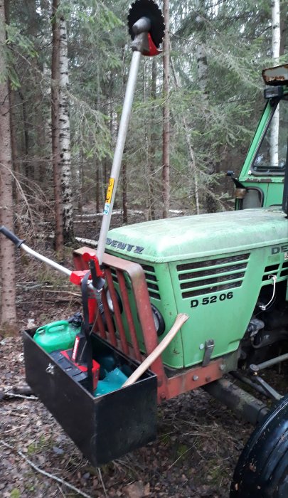 Svetsad ram med verktygslåda på traktor innehållande motorsåg, yxa och bensindunk i skogen.