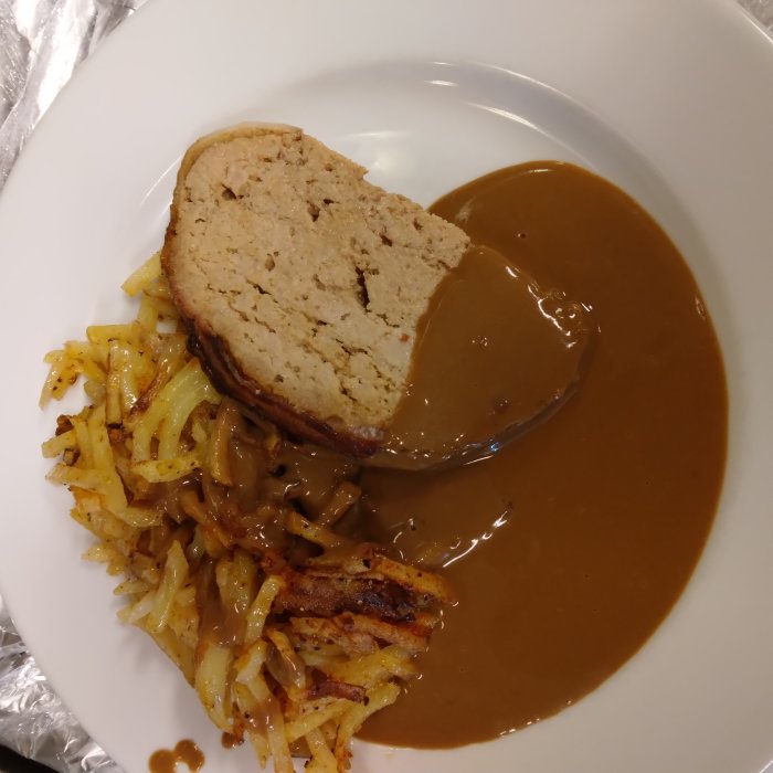 Skiva av baconlindad köttfärslimpa med gräddsås och råstekt potatis på en vit tallrik.