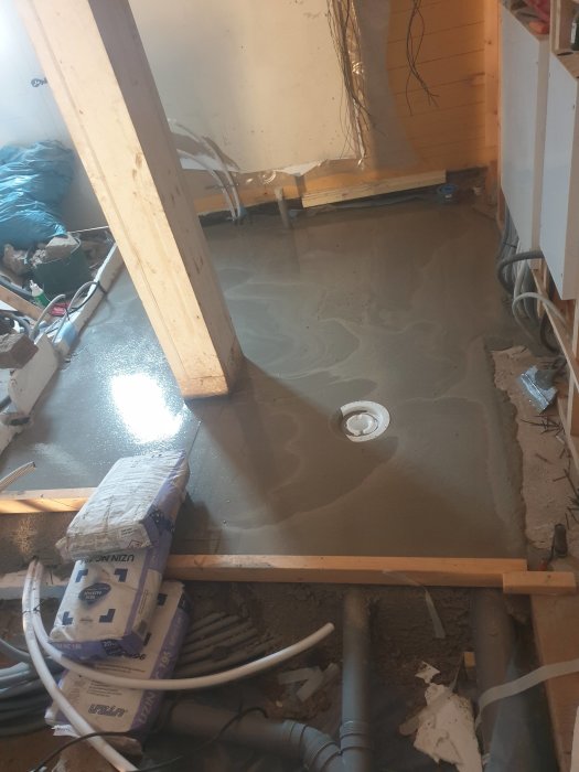 Grovspacklat badrumsgolv med synliga rör och kablar, och byggmaterial spridda ovanpå.