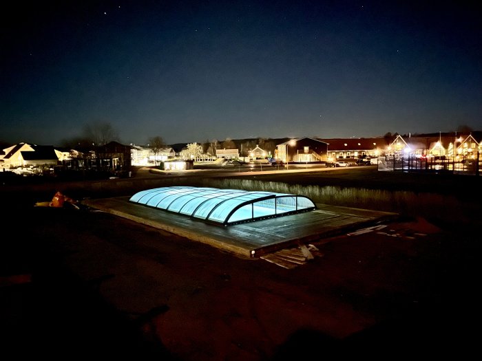 Kvällsbild av en pool med övertäckning nära bebyggelse, belysning och stjärnhimmel syns.