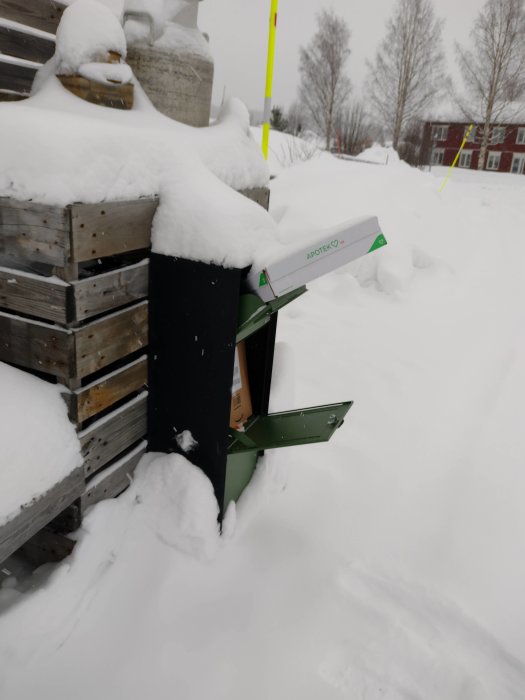 En överfylld grön postlåda delvis nedsnöad med paket som sticker ut i ett snöigt landskap.