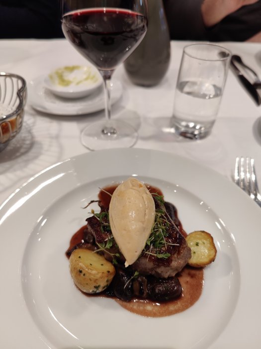 Kalvfilé serverad med ankeleverskum, svamp och potatis på en vit tallrik med rödvinsglas i bakgrunden.