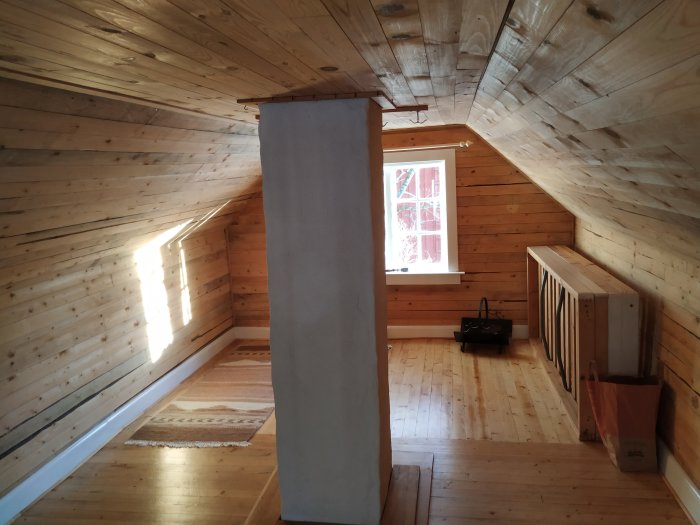 Inredning i en stuga med träpaneler på väggar och tak, synligt skorstensrör och trägolv med en matta.