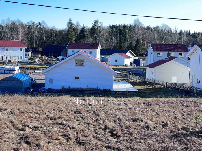 Nybyggt staket längs med vitmålat hus mot en väg och grannområde, med obebyggd naturområde i förgrunden.