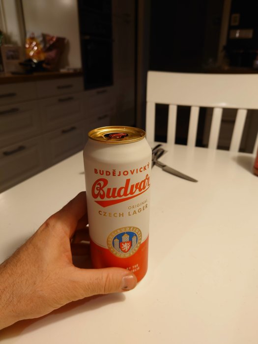 Hand håller en ölburk av märket Budweiser Budvar, tjeckisk lager, på ett vitt köksbord.