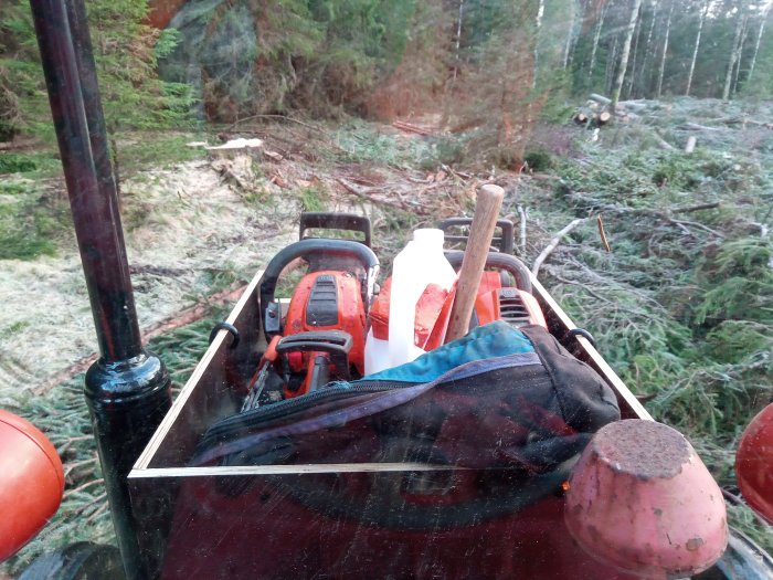 Vy genom hyttfönstret på en skogsmaskin med en röjsåg, verktyg och skogslandskap i bakgrunden.