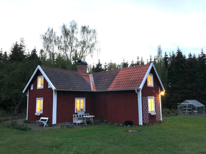 Ett traditionellt rött hus med vita knutar i skymningen, belyst fönster, gräsmatta och växthus.