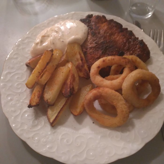 Klyftpotatis, griljerad fläskytterfile, örtagårdssås och lökringar på en vit tallrik.
