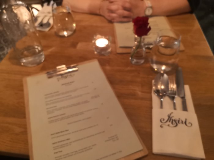 Otydlig bild av restaurangbord med menyer, vattenglas, ljus och en röd ros i fokus.