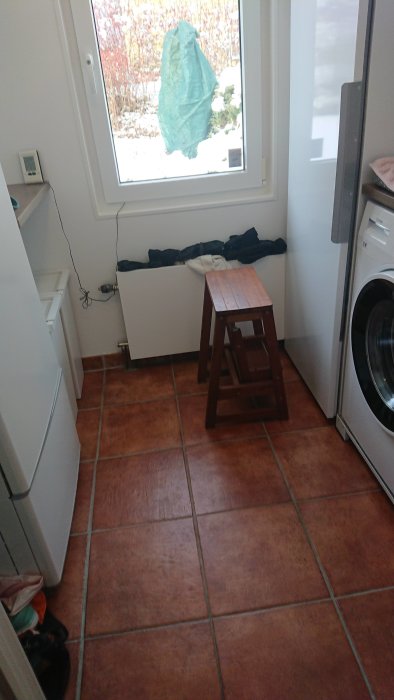 En tvättstuga med tvättmaskin, pall och elektronisk utrustning nära en fönsterbräda och en yttervägg.