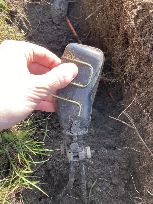 Hand håller en svart plastdosa med kablar i en grävd grop under markarbete.