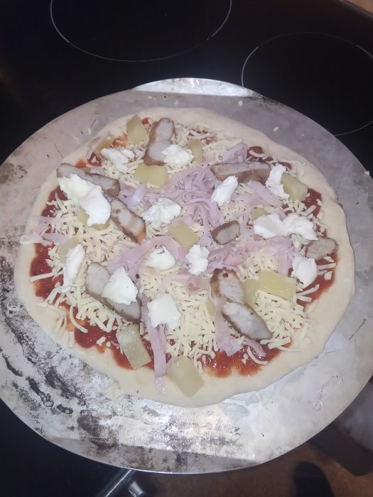 Olagad pizza med riven ost, skinka, ananas och kyckling på en baksten.