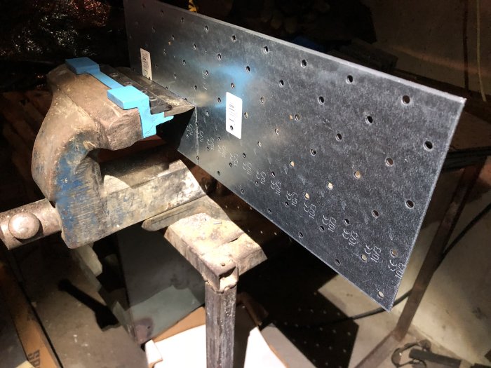 Metal plate with multiple holes clamped in a vise, used for making custom brackets.