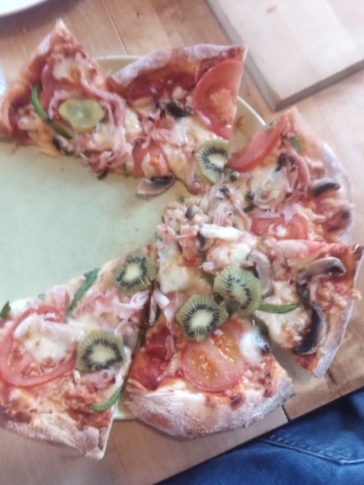 Pizza med olika toppings inklusive skivor av kiwi, tomater och skinka på en tallrik.